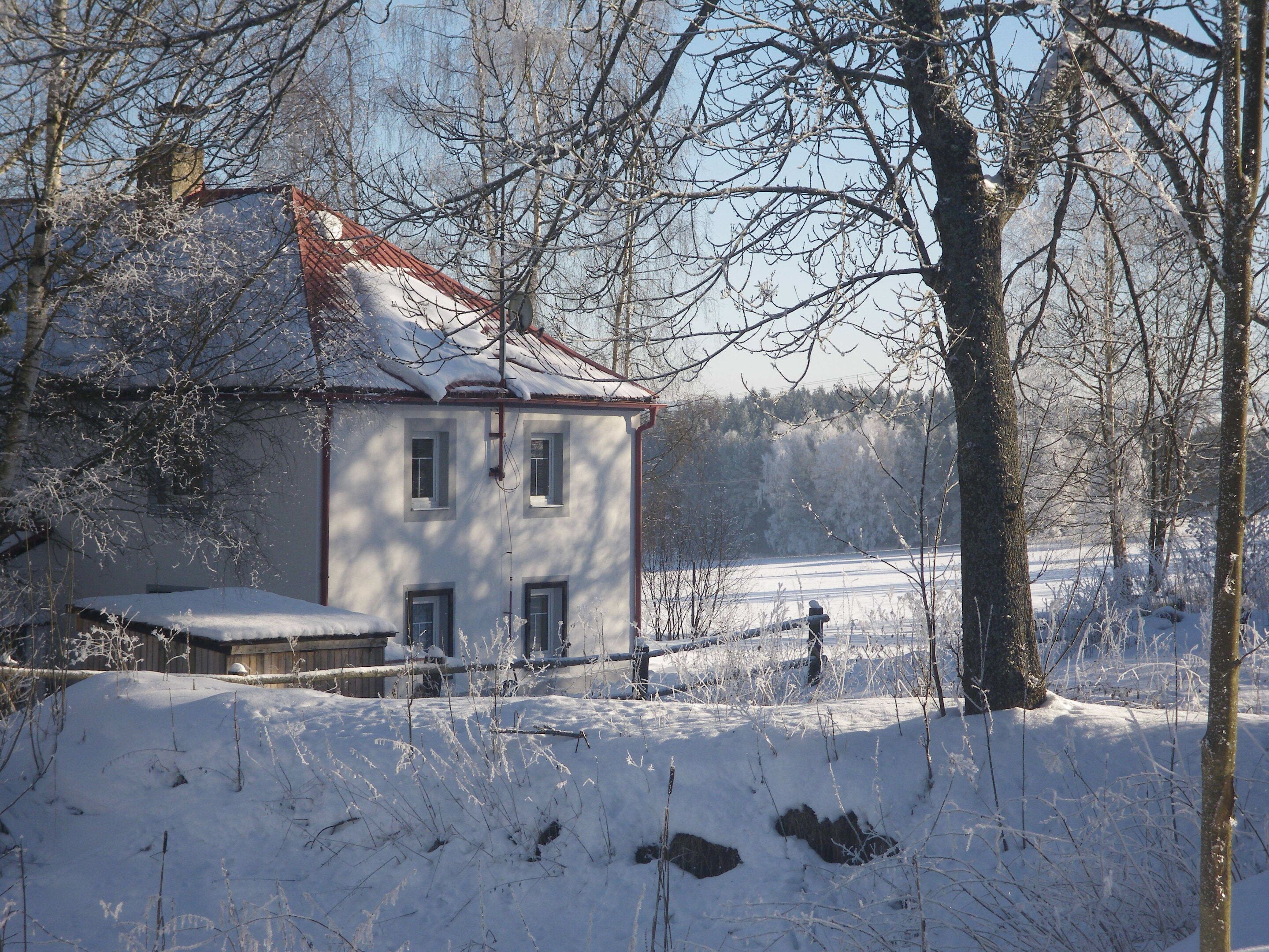 Une maison à deux étages, avec un toit rouge, entourée d’un paysage enneigé et d’arbres.