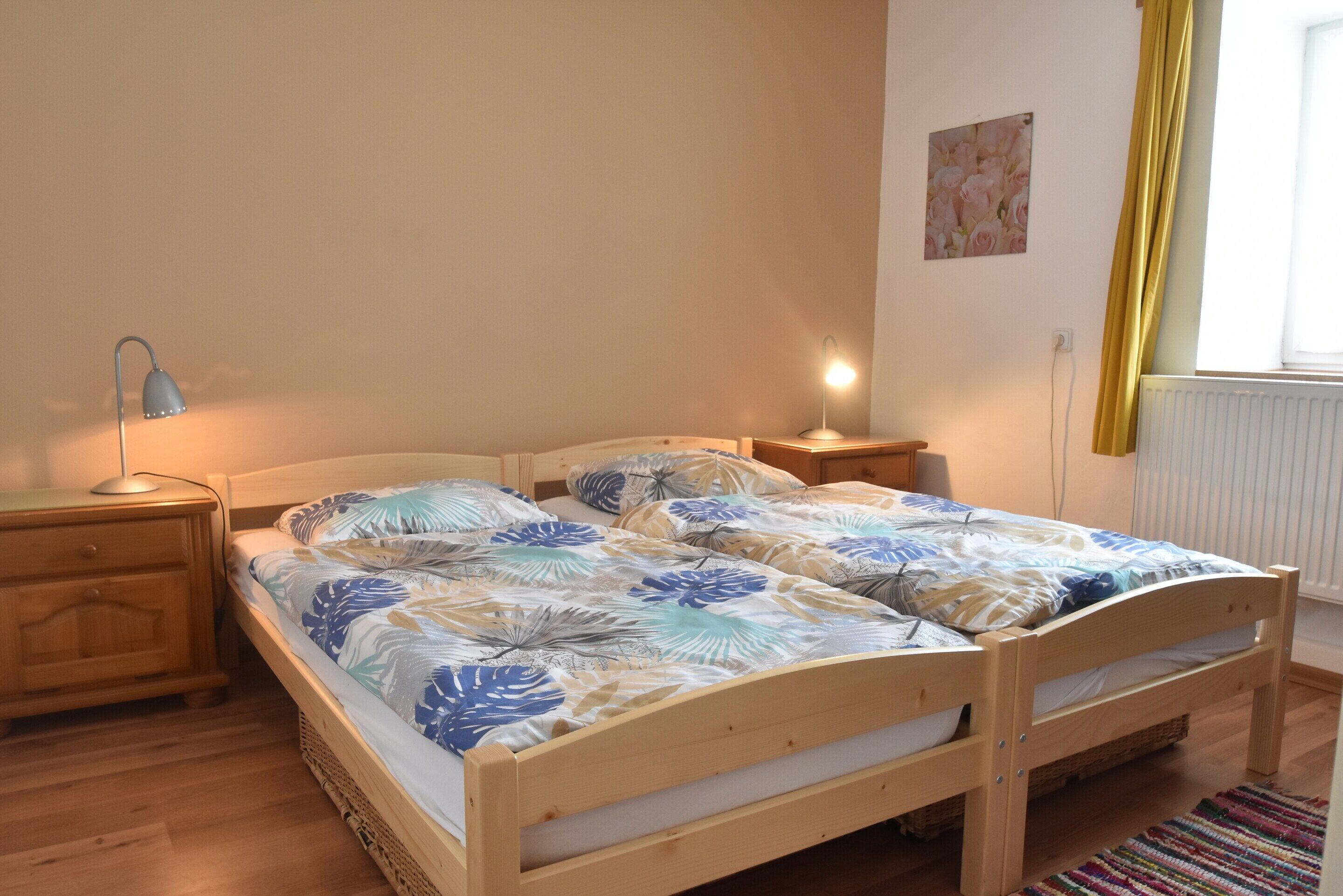 A wooden bed with a floral bedspread, a bedside lamp, a nightstand, and a window with curtains.