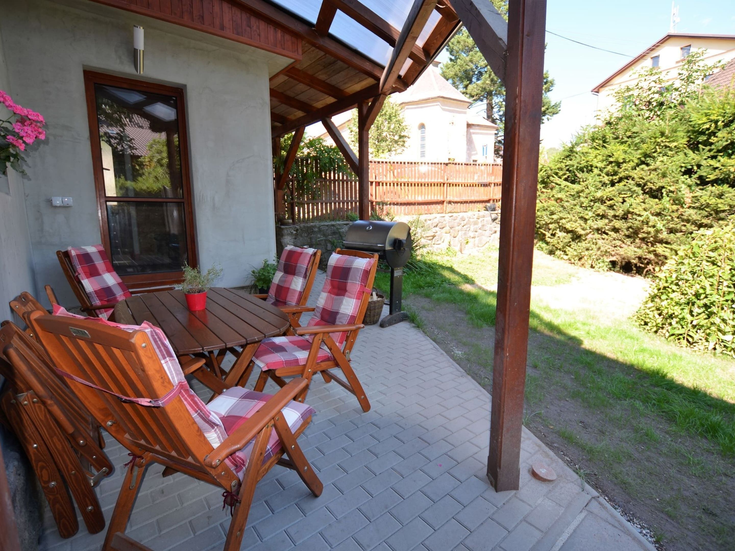 A patio with a wooden table and chairs, a barbecue grill, and a view of a church and fence in the background.