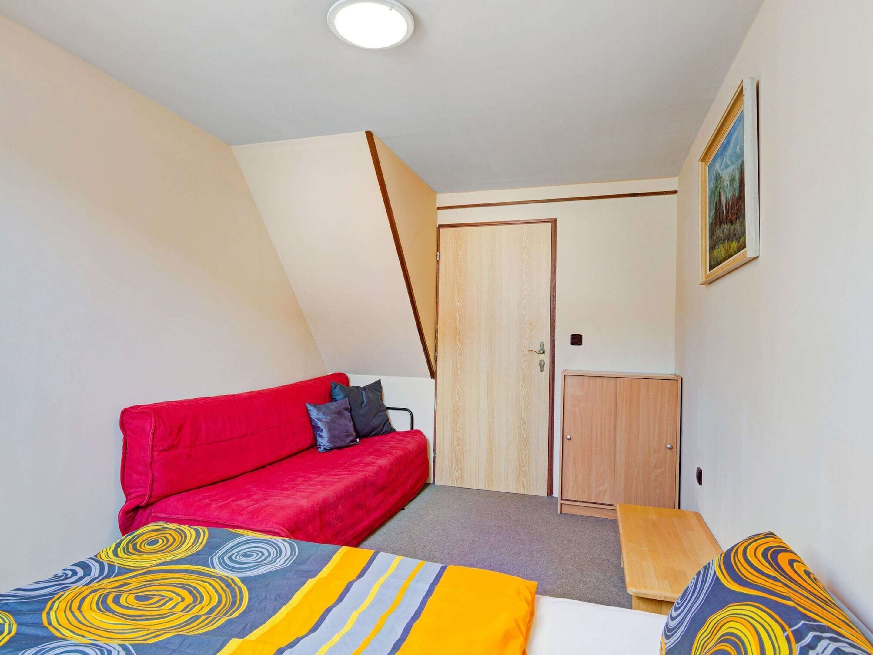A small, well-lit room with a red sofa, a wooden wardrobe, and a painting on the wall.