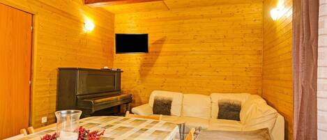 A cozy cabin room with a wooden interior, a television mounted on the wall, a sofa, a dining table with a checkered tablecloth, and a glass vase with red berries.