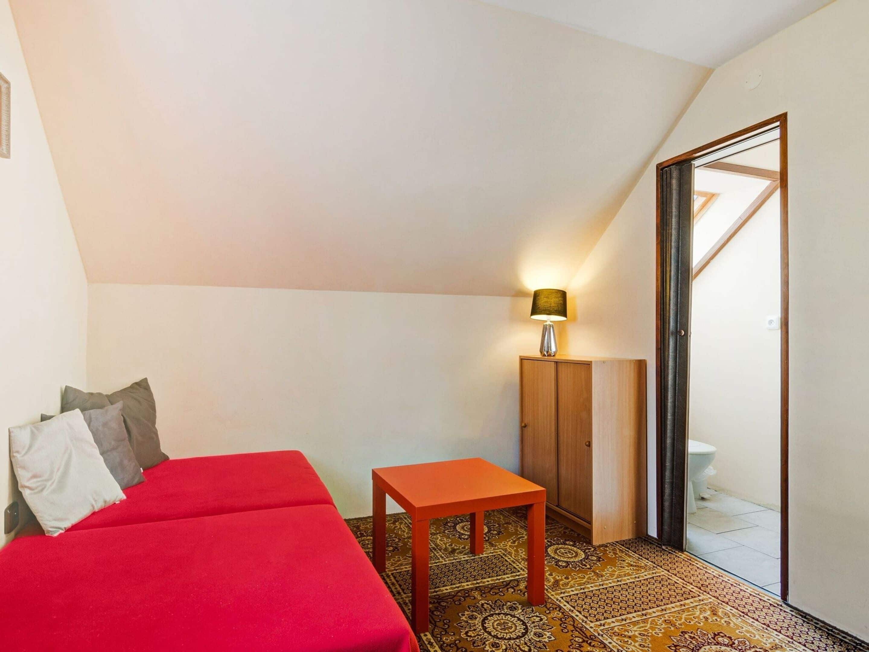 A small, cozy room with a red sofa, a wooden cabinet, and a small table. There is a lamp on the cabinet, and a door leading to a bathroom.