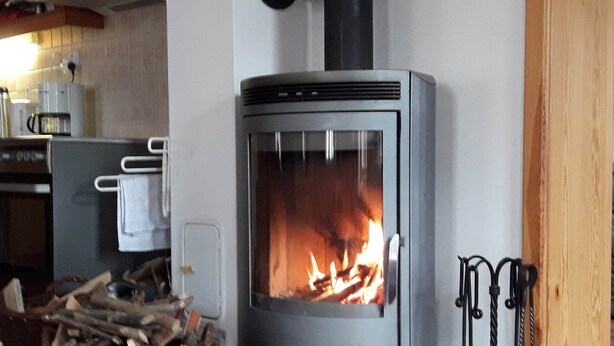 A wood-burning stove with a glass door, a basket of firewood, and a kitchen area with a coffee maker and a kettle. - Holiday Home Rudnik Near Ski Lift (Rudník)