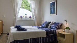 A bedroom with a bed, a nightstand, a bedside lamp, a window with curtains, a radiator, and a painting on the wall.