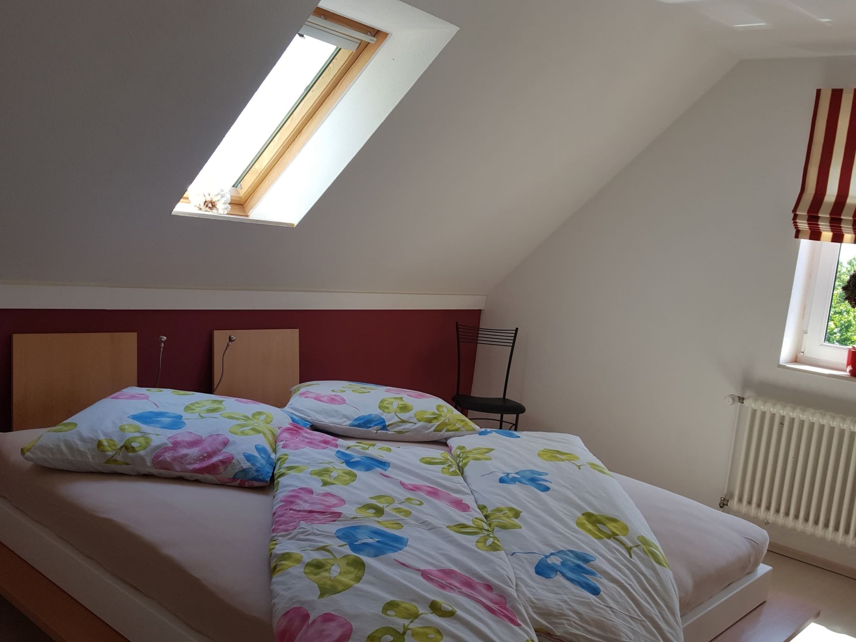 Quarto com teto inclinado, claraboia, radiador e janela com veneziana listrada.