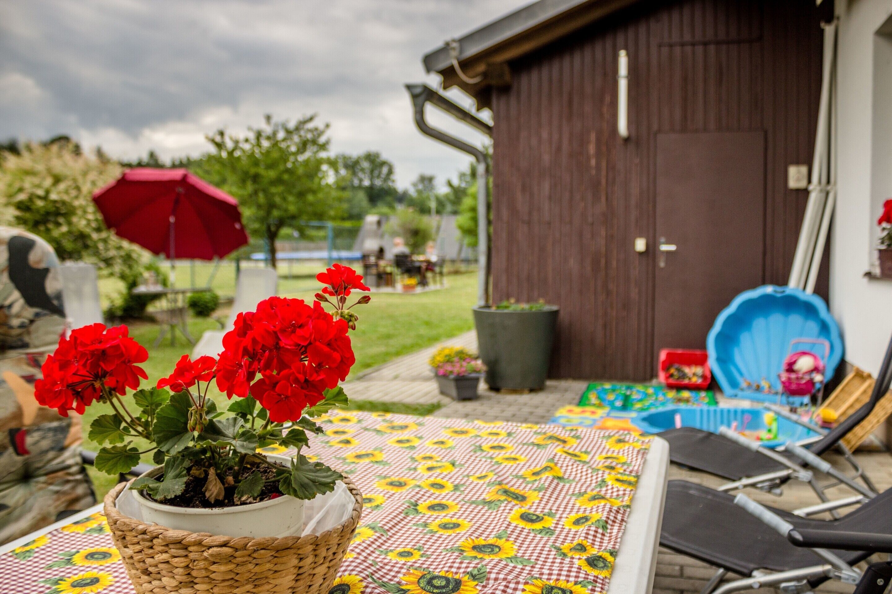 A patio with a table covered in a sunflower-patterned tablecloth, a wicker basket with red flowers, and a red umbrella in the background.
