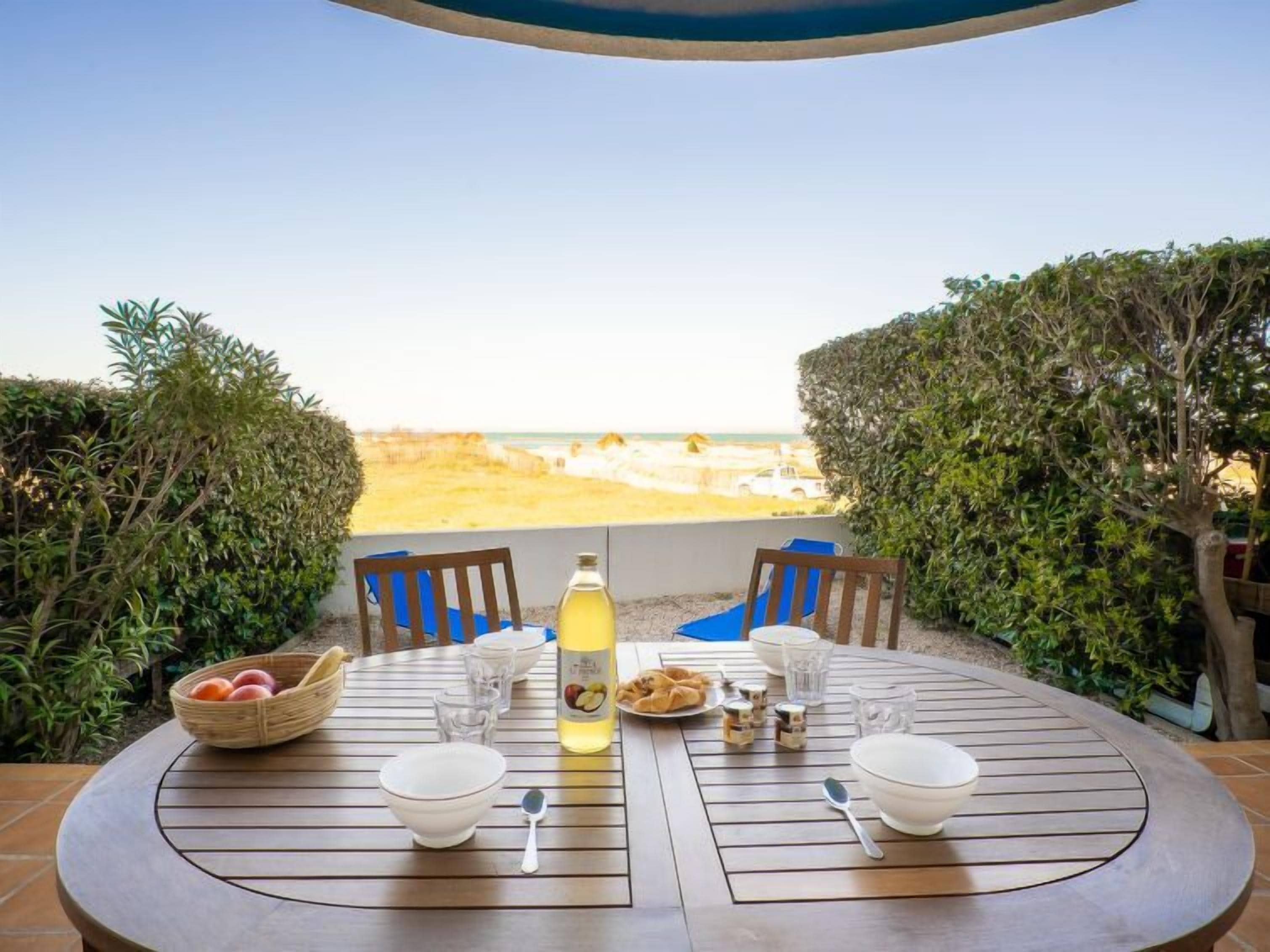 Een ronde houten tafel met stoelen, een fles drank en een bord eten, met uitzicht op een strand.