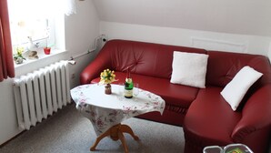 A red leather sofa with white cushions, a small round table with a floral tablecloth, a radiator, and a window with curtains. - Restful Apartment in Neukloster Germany near Lake (Neukloster)