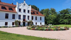 A large white building with a red-tiled roof, multiple windows, and a central entrance with a clock. - Wohnung in Satow mit Garten (Satow)