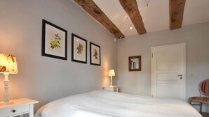 Ein Schlafzimmer mit einem großen Bett, zwei Nachttischen mit Lampen, gerahmten Pflanzenbildern an der Wand, einer Tür und freiliegenden Holzbalken.