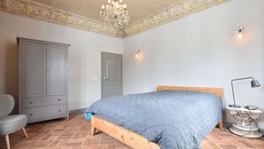 Ein Schlafzimmer mit einem hölzernen Bett, einem grauen Kleiderschrank, einem grauen Sessel, einer Deckenleuchte und einem Fliesenboden.