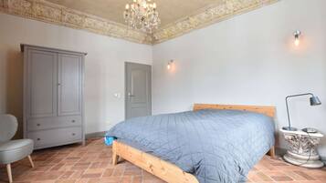 Ein Schlafzimmer mit einem hölzernen Bett, einem grauen Kleiderschrank, einem grauen Sessel, einer Deckenleuchte und einem Fliesenboden.
