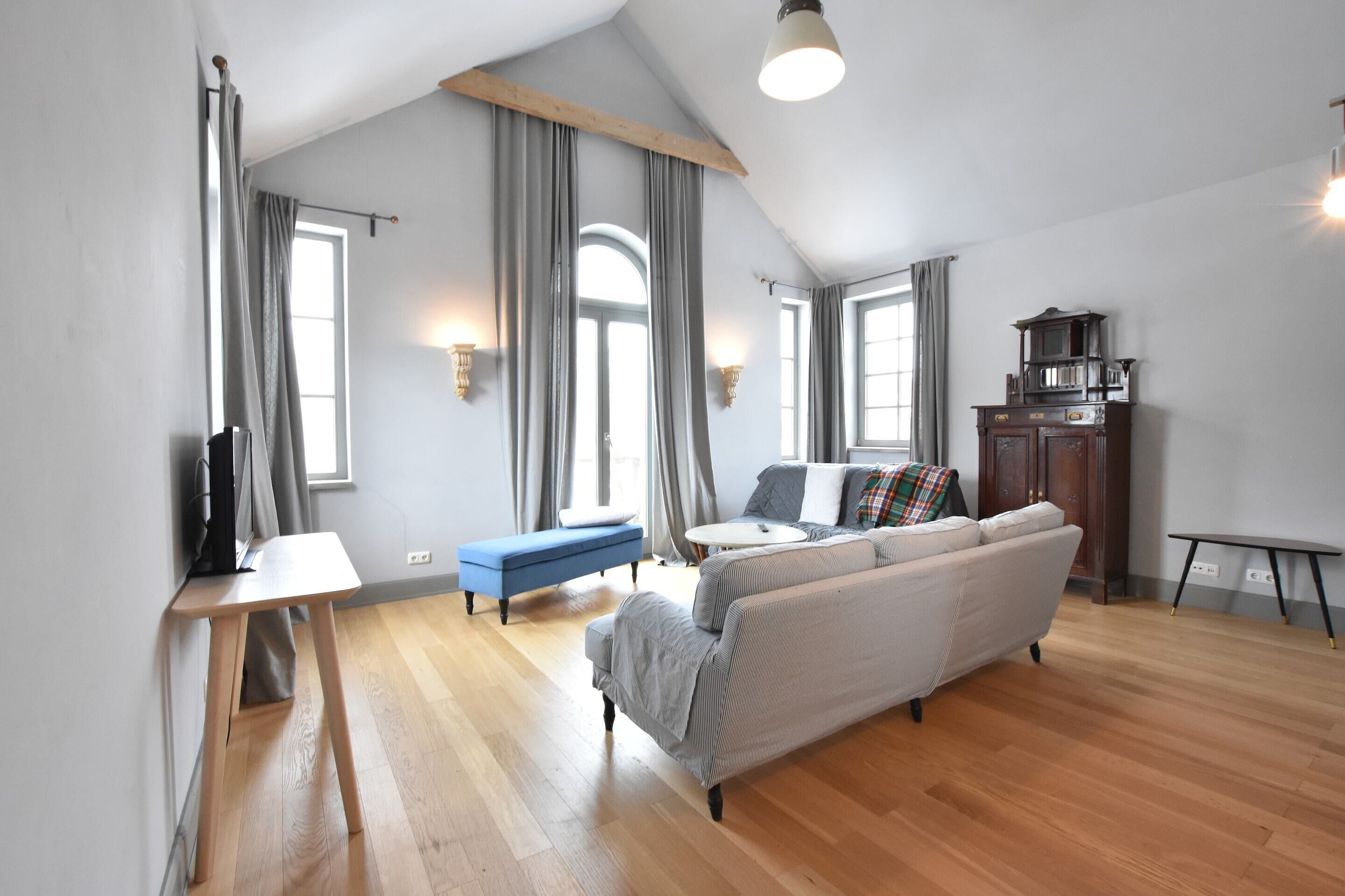 Ein geräumiger Wohnbereich mit hohen Decken, Holzboden, einer Couch, einer blauen Bank und einem Fernseher auf einem Standfuß.