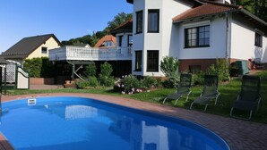 Ein Schwimmbad mit Holzterrasse, ein weißes Haus mit Balkon und ein Garten mit Blumen und Stühlen.