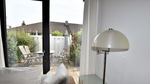 A room with a white sofa, a glass table, and a floor lamp. There is a view of an outdoor patio with a white fence and a table with chairs. - Garten-ferienwohnung in Landlicher Idylle (Boiensdorf)
