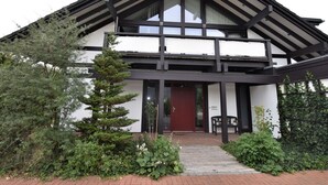 A two-story building with a black and white exterior, a red door, and a small garden area with shrubs and a tree. - Garten-ferienwohnung in Landlicher Idylle (Boiensdorf)