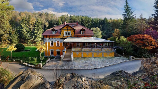 Une grande maison à deux étages, dotée d’un toit à pignon bien visible, entourée d’un jardin luxuriant et bordée par une forêt.