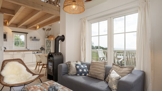 A cozy living room with a sofa, a wicker chair, a table with a rustic tablecloth, and a large window with a view. - Bungalow by the Sea With Sauna in Germany (Sierksdorf)