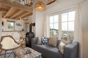 A cozy living room with a sofa, a wicker chair, a table with a rustic tablecloth, and a large window with a view. - Bungalow by the Sea With Sauna in Germany (Sierksdorf)