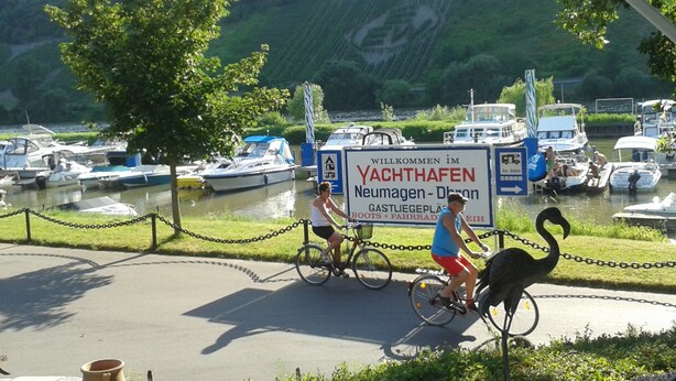 A signboard with text welcoming visitors to a location, a body of water with boats, and a cyclist on the road. - Modern Holiday Apartment With Large Garden Near the River Moselle (Neumagen-Dhron)