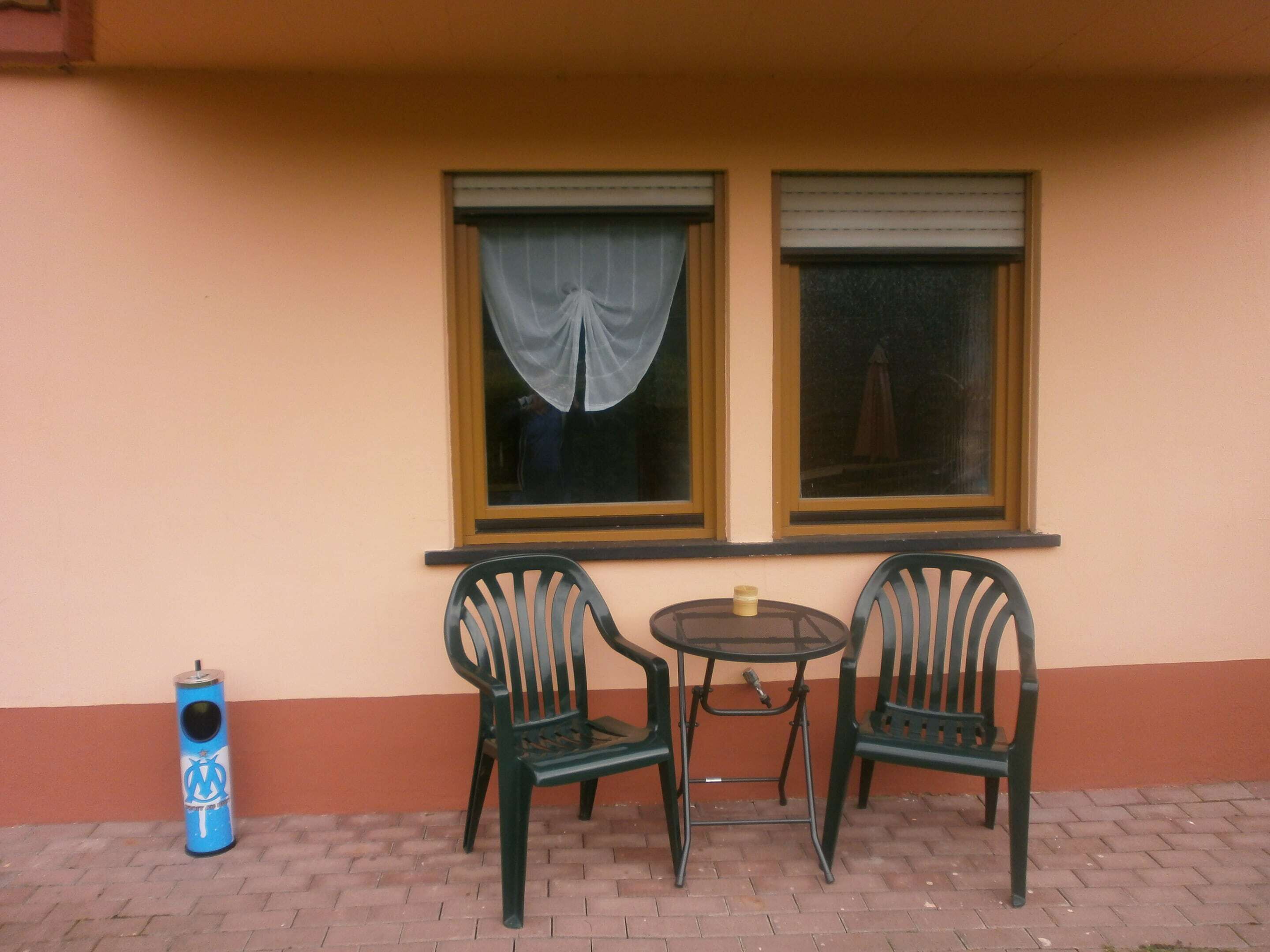 A small outdoor seating area with two chairs and a table, a trash bin, and two windows with curtains.