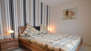 A neatly made bed with a plaid bedspread and two pillows, flanked by bedside tables with lamps, in a room with striped wallpaper and a single wall-mounted painting. - Modern Holiday Apartment With Large Garden Near the River Moselle (Neumagen-Dhron)