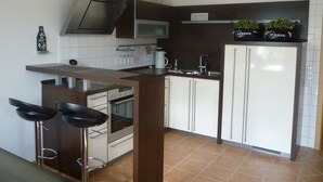 Eine moderne Küche mit Einbauofen, Mikrowelle, Kaffeemaschine und Kühlschrank. Zwei Barhocker und ein Regal mit Dekorationselementen.