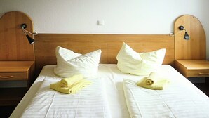Ein Hotelzimmer mit hölzernem Kopfteil, zwei weißen Kissen, einem ordentlich bezogenen Bett mit weißer Bettwäsche und einem Nachttisch mit Lampe.
