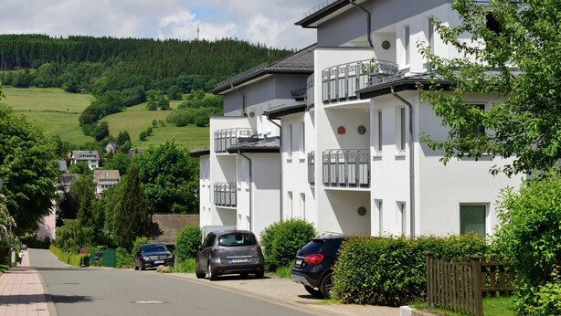 Ein Wohngebäude mit Balkonen, eine gepflasterte Straße und eine hügelige Landschaft im Hintergrund.