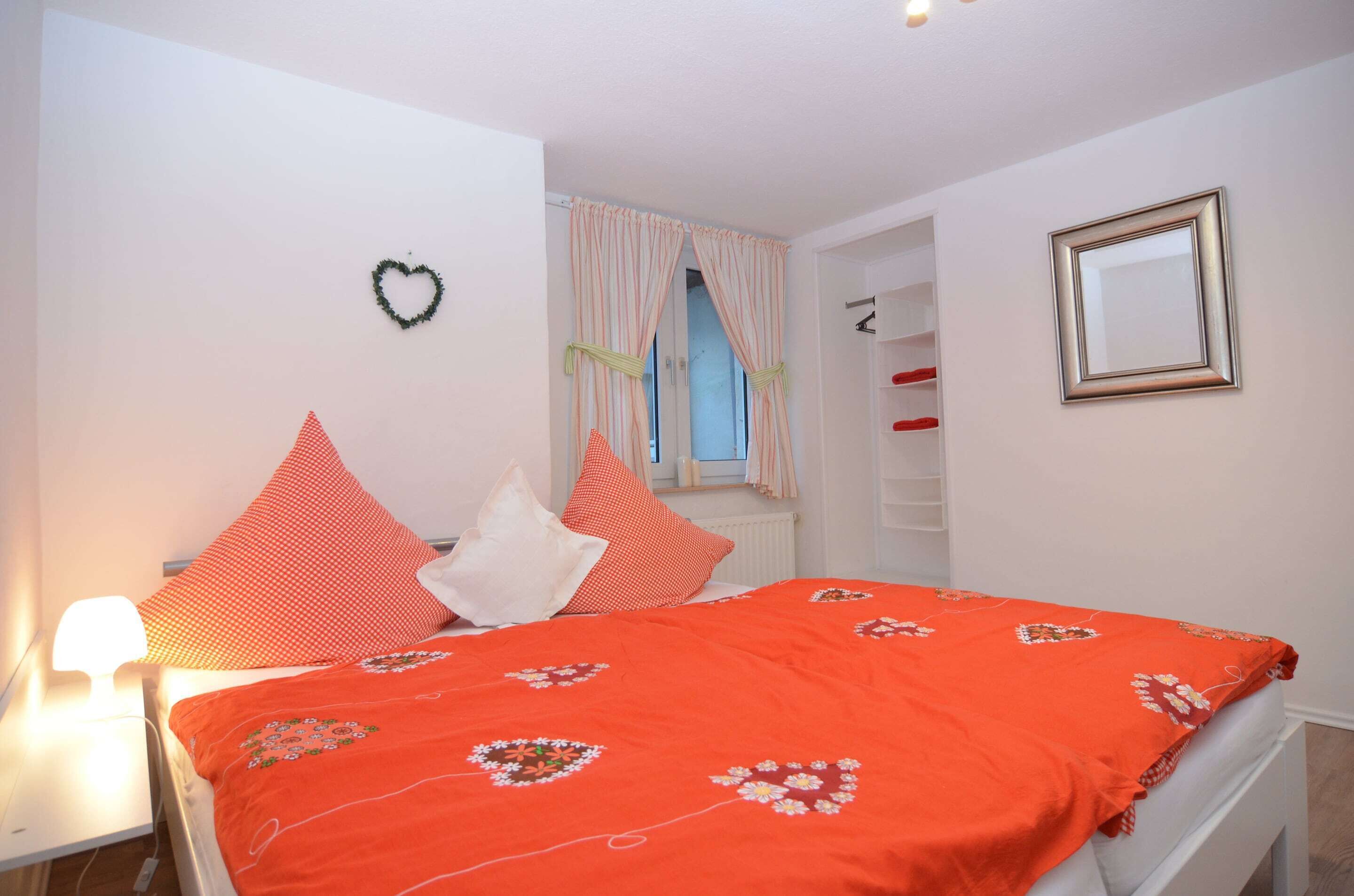 Een slaapkamer met een bed, kussens, een hartvormige decoratie, een raam met gordijnen, een spiegel en een plank met rode objecten.
