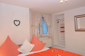 Ein Schlafzimmer mit Bett, Kissen, einem Fenster mit Vorhängen, einem Heizkörper, einem Kleiderschrank und einem Spiegel.