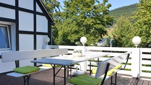 A wooden deck with a white table and benches, a white fence, and a view of trees and hills.