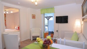 A room with a bed, a white armchair, a dining table with a fruit bowl, a television, and a window with a view of greenery.