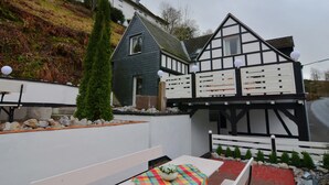 Uma casa tradicional em estilo enxaimel, com telhado inclinado, um pequeno jardim com um banco e uma mesa com toalha colorida.