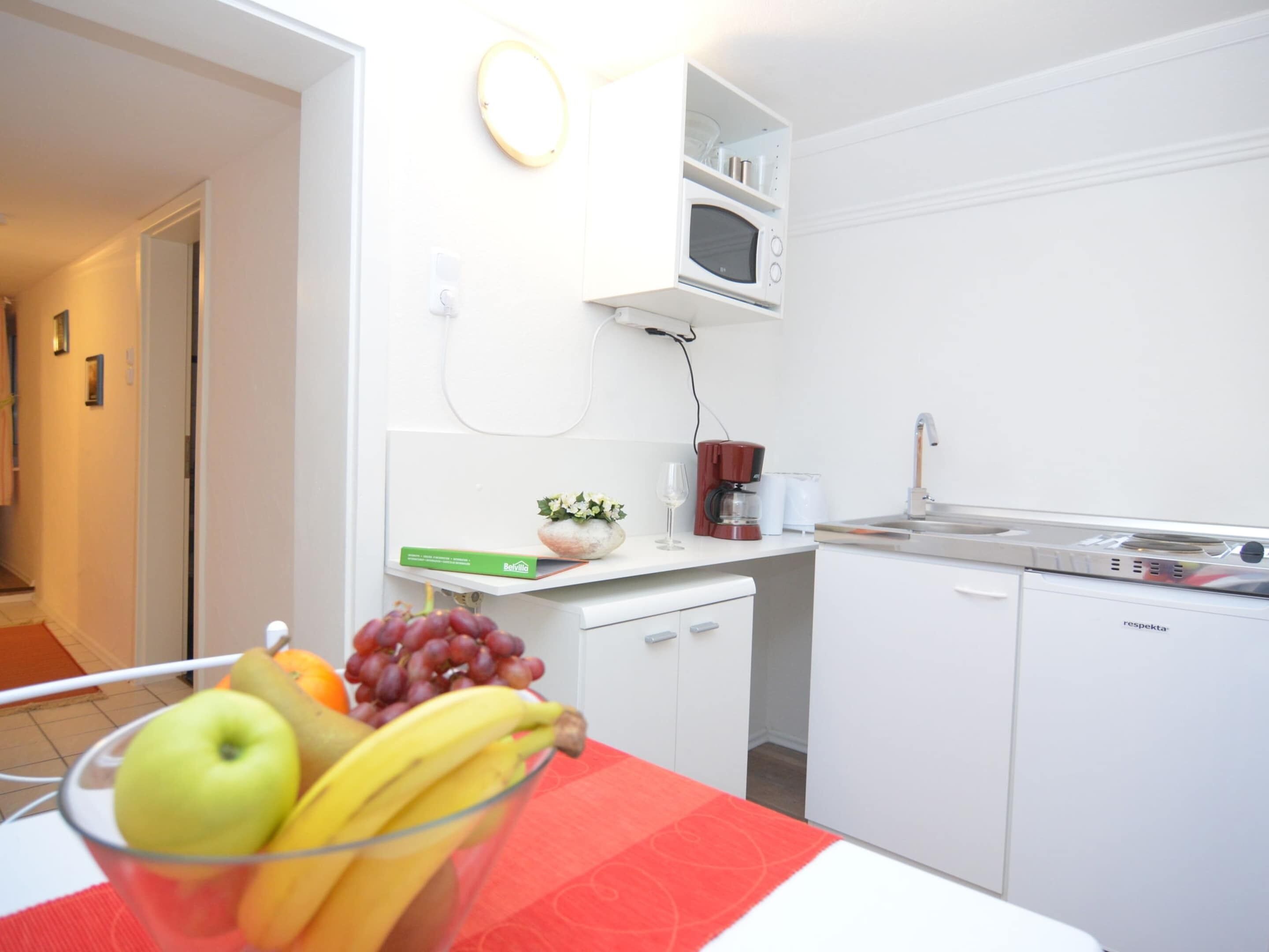Uma cozinha moderna com armários brancos, micro-ondas e cafeteira. Há uma tigela de fruta na bancada.