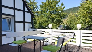 Una terraza de madera con una mesa y bancos blancos, rodeada por una valla blanca y árboles.