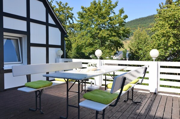 Une terrasse en bois avec une table et des bancs blancs, entourée d’une clôture blanche et d’arbres.