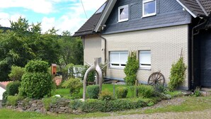 Una casa de dos pisos con techo inclinado, fachada de ladrillo y un pequeño jardín con muro de piedra y diversas plantas.
