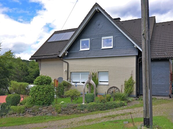 Une maison à deux étages avec un toit en pente, des panneaux solaires et un jardin bordé d’un mur en pierre.