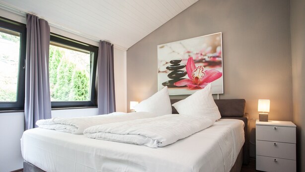 A double bed with white linens, a nightstand with a lamp, and a painting of flowers on the wall. - Stylish Holiday Home near Winterberg with Private Sauna House, Terrace, & Garden (Winterberg)