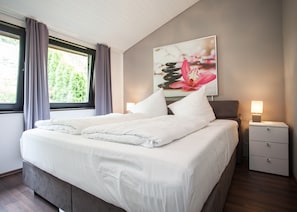 A double bed with white linens, a nightstand with a lamp, and a painting of flowers on the wall. - Stylish Holiday Home near Winterberg with Private Sauna House, Terrace, & Garden (Winterberg)