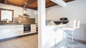 A modern kitchen with white cabinets, a built-in oven, and a wooden ceiling. There is a bar area with two white bar stools and a clock on the wall. - Stylish Holiday Home near Winterberg with Private Sauna House, Terrace, & Garden (Winterberg)