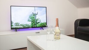A room with a flat-screen TV mounted on a white wall, a table with a bottle of champagne and two glasses, and a black chair. - Stylish Holiday Home near Winterberg with Private Sauna House, Terrace, & Garden (Winterberg)