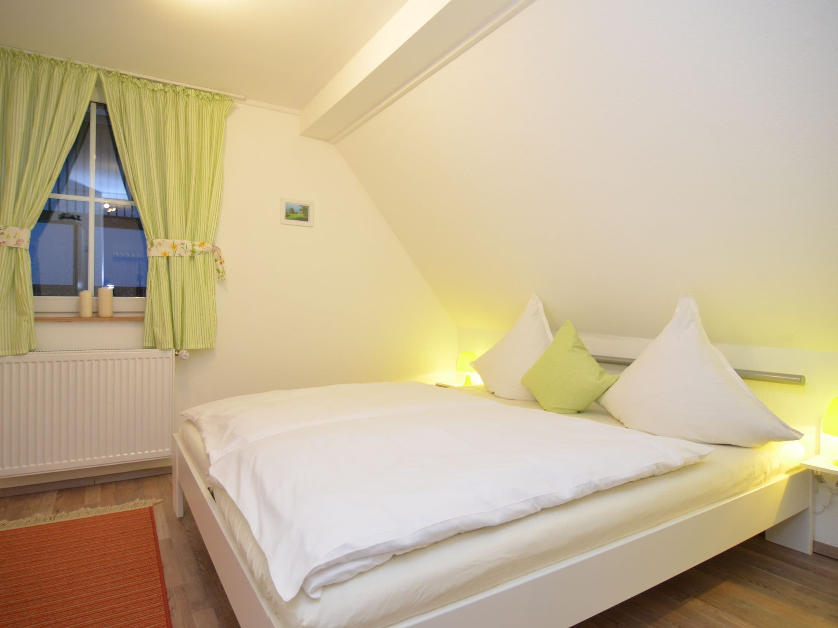 A bedroom with a single bed, a window with curtains, a radiator, and a nightstand with a lamp.