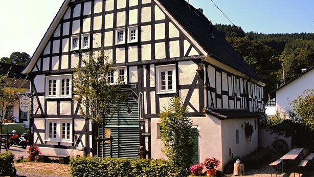 A traditional half-timbered building with a steep gabled roof, a green door, and a small garden area with benches and potted plants. - Altes Ferienhaus mit Sauna in Heinsberg (Kirchhundem)