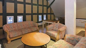 A cozy attic room with exposed wooden beams, a patterned sofa, a round table, and a view of the outside through a window. - Altes Ferienhaus mit Sauna in Heinsberg (Kirchhundem)