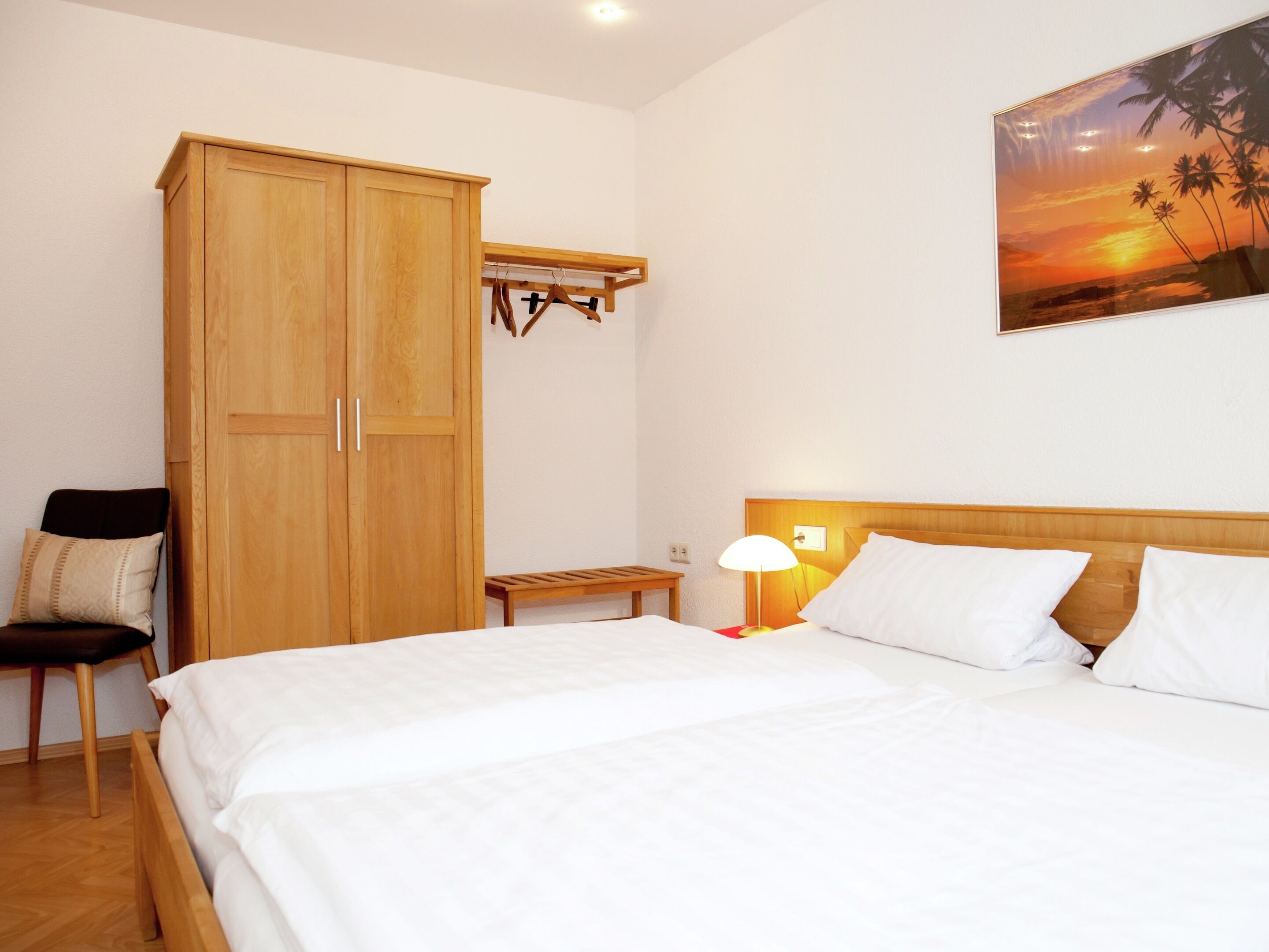 A bedroom with a large wooden wardrobe, a bench, a bedside table with a lamp, and a bed with white bedding.