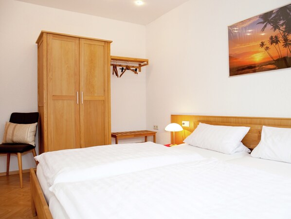 Une chambre avec une grande armoire en bois, un banc, une table de chevet avec une lampe et un lit avec une literie blanche.