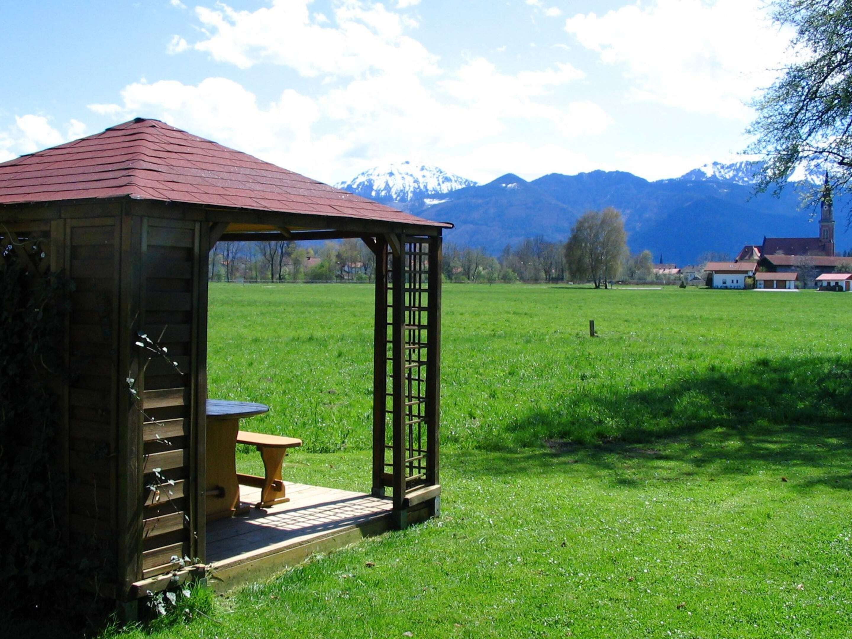 A wooden gazebo with a bench, set in a grassy field with a mountain backdrop.