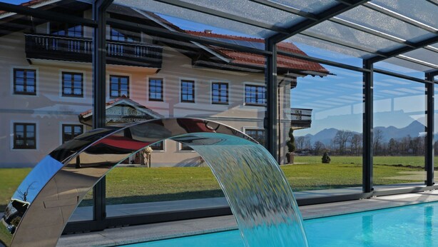 Piscina com um moderno detalhe aquático, área coberta com teto de vidro e um edifício com telhado de telhas vermelhas ao fundo.
